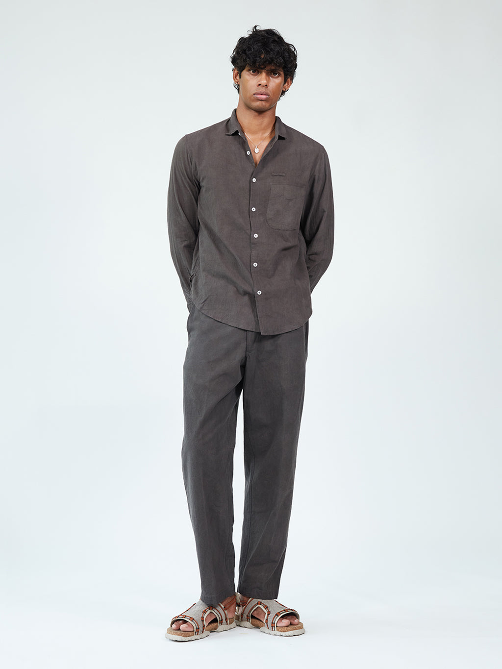 A man modeling a dark brown linen two-piece set consisting of a long-sleeve button-up shirt and matching trousers, paired with brown leather slide sandals, against a plain white studio background.
