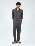 A man modeling a dark brown linen two-piece set consisting of a long-sleeve button-up shirt and matching trousers, paired with brown leather slide sandals, against a plain white studio background.